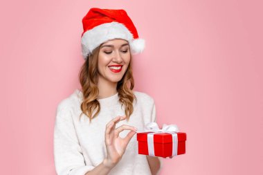Noel Baba şapkalı, gülümseyen ve elinde pembe stüdyo arka planında izole edilmiş kırmızı hediye kutusu tutan beyaz bir kadın. Uzayı kopyala Yeni yıl hediyeleri konsepti