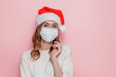 Koruyucu tıbbi maske takan genç esmer bir kadın. Bir noel şapkası ve pembe stüdyo arka planında poz veren beyaz bir kazak.