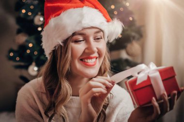 Yılbaşı tatili konsepti. Noel şapkalı, gülümseyen beyaz bir kızın portresi. Kırmızı bir hediye kutusu açıyor. Bir çelenk ile bir Noel ağacının arka planında.