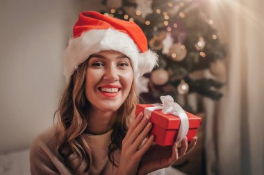 Beyaz kazak ve Noel Baba şapkalı genç bir kadın kırmızı bir hediye kutusu tutuyor, gülümsüyor ve kameraya bakıyor. Koronavirüs salgınının karantinası sırasında yeni yılı kutluyoruz.