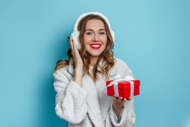 Yapay beyaz kürk giyen, gülümseyen, elinde kırmızı bir hediye kutusu tutan ve mavi arka planda izole edilmiş kameraya gösteren genç ve mutlu bir kadın. Yeni yıl hediyeleri konsepti