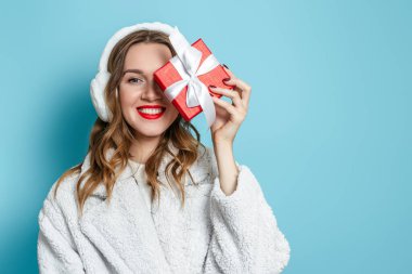 2021 yılbaşı hediyeleri. Beyaz kürklü ve kürklü genç mutlu bir kadın hediye kutusuyla gözlerini kapatıyor ve mavi bir stüdyo arka planında izole bir şekilde gülümsüyor. Boşluğu kopyala