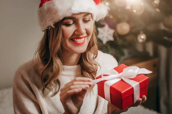 Happy woman in a New Year\'s costume smiles and unpacks a gift box against the background of New Year\'s decorations and a Christmas tree with garlands