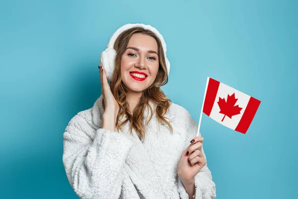 快乐的高加索女人 身穿假的白毛皮外套 微笑着 拿着一面小加拿大国旗 与蓝色的工作室背景隔离在一起 — 图库照片
