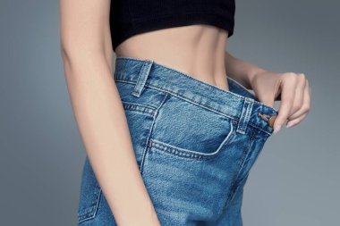 Gri arka planda izole edilmiş, büyük kot pantolonlu zayıf bir kadın cesedi. Kilo verme kavramı. Kız elinde kot pantolon tutuyor ve ince bir bel gösteriyor. Büyük beden kotlu kadın, diyet konsepti