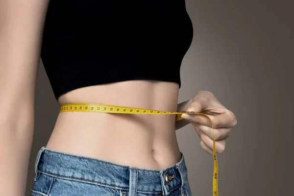 A young girl in a black top and blue jeans measures her waist with a centimeter on a dark gray background, copy space. Female body, slim waist. The girl\'s healthy athletic body, diet, weight loss, calorie count. Slimming concept