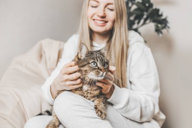 Kedi, genç sarışın beyaz bir kadının bacaklarının üzerinde uzanmış onu okşuyor ve gülümsüyor.