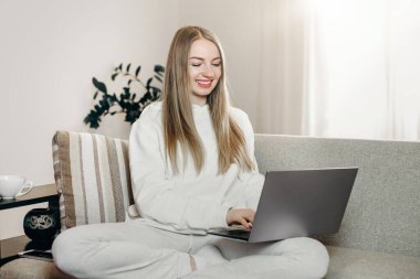 Genç sarışın kadın evinde kanepede oturuyor dizüstü bilgisayarla gülümsüyor ve koronavirüs salgını sırasında karantinada çalışıyor. Kadın, bilgisayarda kapüşonlu bir kapüşonluyla evde oturuyor.