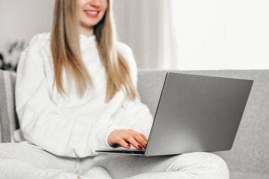 Genç sarışın bir kadın kanepede oturuyor ve laptopunu kullanıyor. Uzaktan eğitim. İnternette çalışıyorum. Serbest Çalışan Kız