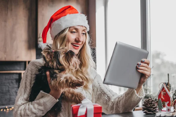 Christmas video calling concept. Young smiling blonde caucasian girl holding a small dog in her arms and making video calls via a tablet, accepts Happy New Year greetings and rejoices