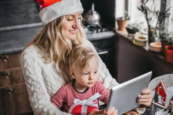 Young Caucasian Mother in New Year\'s Santa hat with her little son are sitting at a table in the kitchen and making video conference calls via a tablet. New year video calls concept