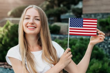 Genç kız gülümsüyor ve küçük bir Amerikan bayrağı gösteriyor ve üniversitenin arka planında duruyor.