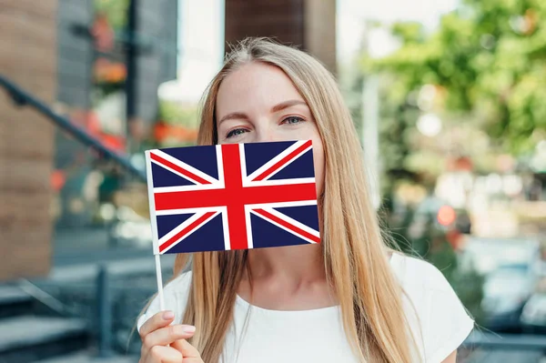 Education abroad. Student girl covers her face with a small UK British England flag and stands near the university on the street
