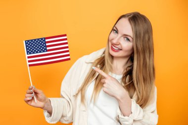 Mutlu sarışın kadın, küçük bir Amerikan bayrağını işaret ediyor ve turuncu stüdyo arka planında izole edilmiş bir şekilde gülümsüyor, elinde ABD bayrağı tutan bir kız, 4 Temmuz Bağımsızlık Günü.