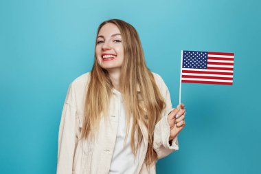 Komik sarışın öğrenci elinde küçük bir Amerikan bayrağı, mavi arka planda izole edilmiş bir gülümseme, elinde ABD bayrağı, 4 Temmuz bağımsızlık günü, kopyalama alanı
