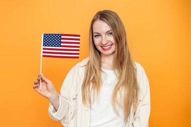 Neşeli genç sarışın kız öğrenci elinde küçük bir Amerikan bayrağı, turuncu arka planda saklanmış bir gülümseme, Amerikan bayrağı taşıyan bir kız, 4 Temmuz Bağımsızlık Günü, kopyalama alanı