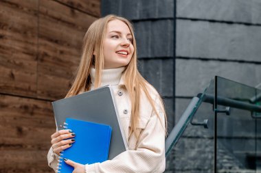 Öğrenci kız dizüstü bilgisayarları elinde tutuyor gülümsüyor, modern bir üniversite binasının arka planına bakıyor.