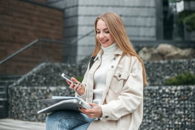 Üniversite binasının arka planında akıllı telefon kullanan, gülümseyen ve kitap okuyan mutlu bir öğrenci.