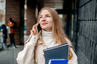 Kafkasyalı sarışın kız cep telefonuyla konuşuyor klasörler, kitaplar, defterler, gülümsemeler, modern bir üniversite binasının arka planına bakıyor.