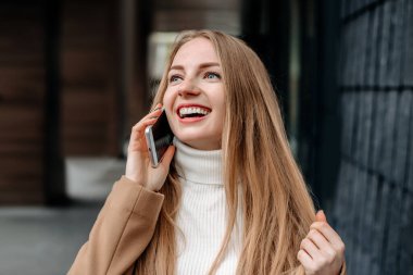 Bej ceketli genç sarışın beyaz kadın cep telefonuyla konuşuyor, gülümsüyor ve bir ofis binasının arka planına bakıyor. Şirket çalışanı. Mutlu kadın bilişim uzmanı.