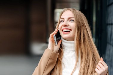 Bej ceketli genç sarışın beyaz kadın cep telefonuyla konuşuyor, gülümsüyor ve bir ofis binasının arka planına bakıyor. Şirket çalışanı. Mutlu kadın bilişim uzmanı. Boşluğu kopyala