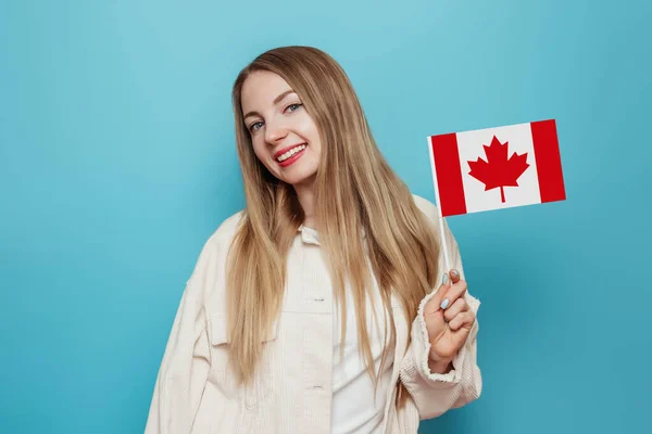喜庆的女生笑着举着小加拿大国旗 望着蓝色背景下的相机 加拿大日 邦联周年纪念日 复制空间 — 图库照片