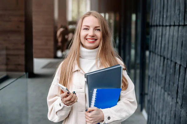 教育概念 白种人的金发女学生手持手机 文件夹 笔记本 看着摄像机 在欧洲大学大楼的背景下微笑着 — 图库照片