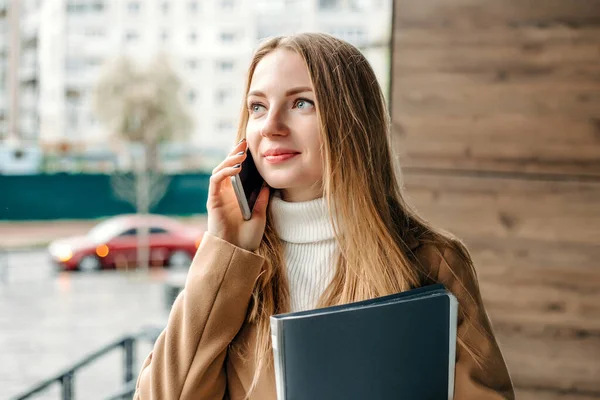 Bej ceketli genç sarışın kadın cep telefonuyla konuşuyor, gülümsüyor, klasörü tutuyor ve bir ofis binasının arka planına bakıyor. Şirket çalışanı. IT uzmanı.