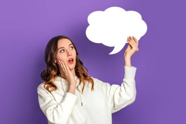 Genç, şok olmuş bir kız öğrenci elinde mor arka planda izole edilmiş boş konuşma balonu kağıdı tutuyor. Yazı için beyaz kağıt bulutu. Uzayı kopyala Model
