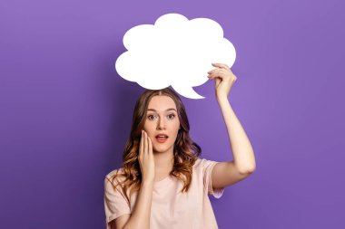 Genç, şaşkın kadının elinde mor arka planda izole edilmiş boş konuşma balonu kağıdı var. Leylak üzerine yazı için beyaz kağıt bulutu. Boşluğu kopyala