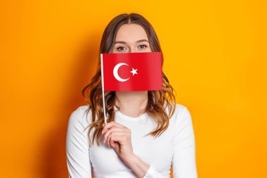 Young woman holding a small turkish flag in her hands and covering her face with it isolated on orange background clipart