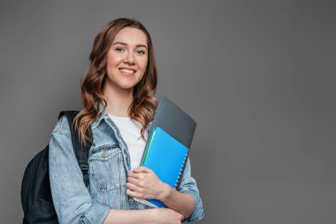 Sırt çantası gülüşlü mutlu kız öğrenci elinde defter, defter, klasör koyu gri arka planda izole edilmiş, kopyalama alanı var. Eğitim, eğitim, sınavlar, İngilizce öğrenme kavramı.