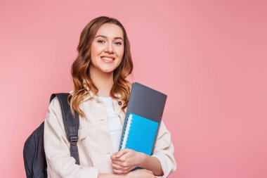 Sırt çantalı, gülümseyen ve elinde bir defter, bir defter, bir defter, pembe arka planda izole edilmiş bir klasör tutan mutlu kız öğrenci, kopyalama alanı. Eğitim, eğitim, sınavlar, İngilizce öğrenme kavramı.