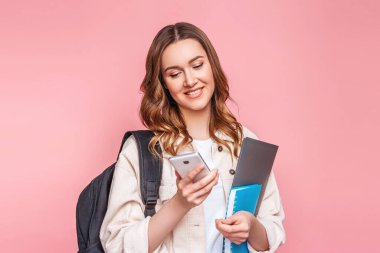 Pembe arka planda izole edilmiş mesajlar yazmak için akıllı telefon kullanan sevimli öğrenci kız. Elinde cep telefonu tutan, gülümseyen ve mesaj okuyan genç mutlu kadın, boşluğu kopyala