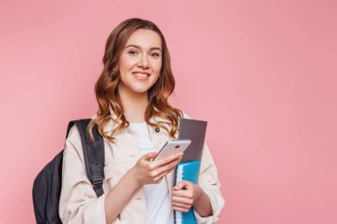 Sırt çantası ve not defteriyle beyaz bir kız öğrenci elinde bir cep telefonu tutuyor ve pembe bir arka planda izole bir şekilde gülümsüyor. Sınavlar, sınavlar eğitim konsepti. Akıllı telefonuyla poz veren genç kadın, boşluğu kopyala