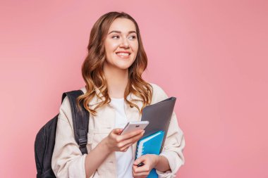 Kız öğrenci gülüyor, başka tarafa bakıyor ve elinde pembe arka planda izole edilmiş bir cep telefonu tutuyor. Sırt çantası, not defteri, not defteri olan genç bir kadın komik mesajlar okuyor, sınav konseptini test ediyor.