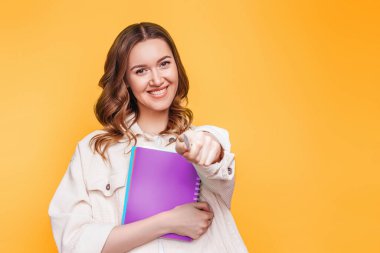Öğrenci kız gülümsüyor ve kamerayı önüne doğrultuyor. Turuncu arka plan üzerinde izole edilmiş, kopyalama alanı. Elinde defteriyle gülen mutlu genç bir kadın, şirket çalışanı.