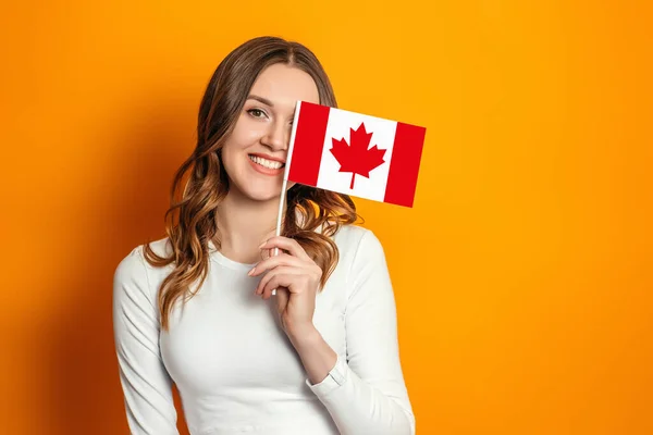student girl cover her face with a small canada flag isolated over orange background, Canada day, holiday, confederation anniversary, copy space