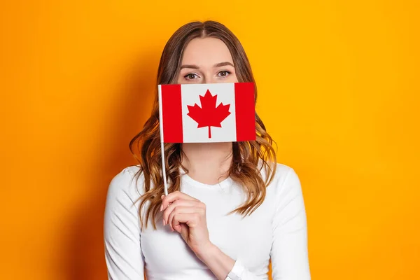 Female student cover her face with a small canada flag isolated over orange background, Canada day, holiday, confederation anniversary, copy space