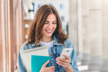 Üniversiteye karşı gülümseyen genç bir kız. Güzel kız öğrenci klasörler, defterler ve cep telefonları tutuyor ve mesajları okuyor. Öğrenme, eğitim kavramı