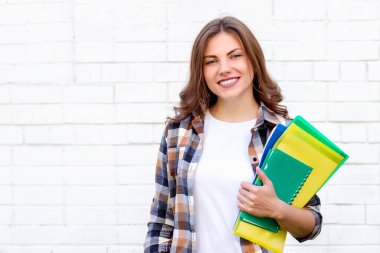 Kız öğrenci ellerinde klasörler ve bir defter tutuyor ve beyaz tuğla bir duvarın arka planında gülümsüyor.