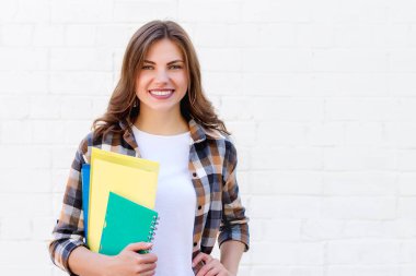 Kız öğrenci ellerinde klasörler ve bir defter tutuyor ve beyaz tuğla bir duvarın arka planında gülümsüyor, kopyalama alanı. Eğitim kavramı