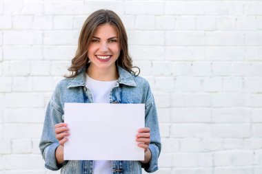 Kız öğrenci gülümsüyor ve beyaz bir duvarın arkasında beyaz bir kağıt tutuyordu.