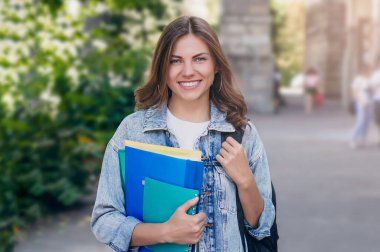 Üniversiteye karşı gülümseyen genç bir kız. Sevimli kız öğrenci klasörleri ve defterleri elinde tutuyor. Öğrenme, eğitim kavramı