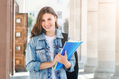 Üniversiteye karşı gülümseyen genç bir kız. Sevimli kız öğrenci klasörleri ve defterleri elinde tutuyor. Öğrenme, eğitim kavramı