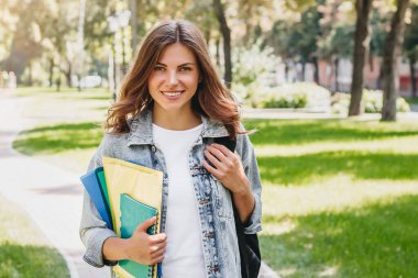 Parkta gülümseyen genç kız öğrenci. Sevimli kız öğrenci elinde klasörler ve defterler tutuyor, kopyalama alanı.