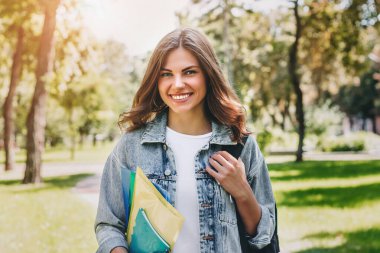 Parkta gülümseyen genç kız öğrenci. Sevimli kız öğrenci klasörleri ve defterleri elinde tutuyor. Öğrenme, eğitim kavramı