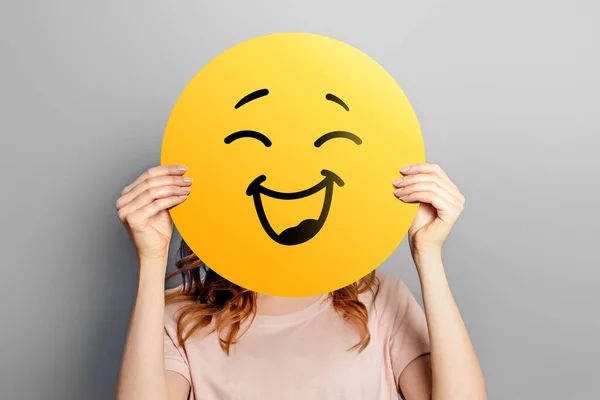 Happy emoji. Girl holds a yellow emoticon with a smile isolated on a gray background. Happy emoji