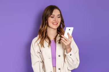 Beyaz tenli bej kadife ceket giyen genç bir kız telefon ekranına bakıyor ve eflatun arka planda sırıtıyor. Boşluğu kopyala