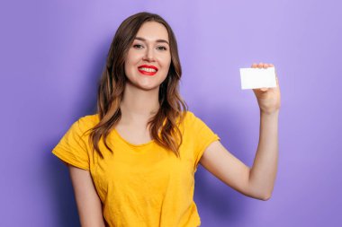 Genç beyaz bir kadın elinde bir kartvizit tutuyor ve gülümsüyor. Stüdyoda leylak bir arka planda sarı tişört giyiyor. Kredi çevrimiçi. Model